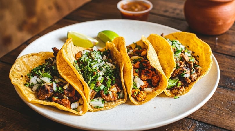 Four street tacos with meat, cilantro, and onions, served with lime wedges and salsa.