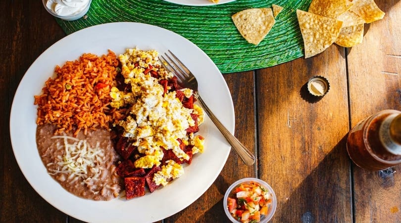 A plate of Mexican breakfast with scrambled eggs, chorizo, rice, and refried beans on a wooden table.